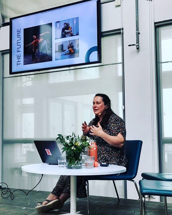 Rosie Heafford sits in front of a screen as she delivers a presentation. On the screen is a picture from We Touch, We Play, We Dance with the caption The Future