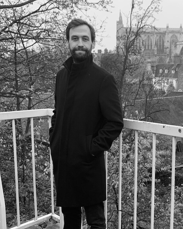 Andrew Bishop, Second Hand Dance Head of Operations with short brown hair, wearing a dark coat, standing with hands in pockets on a balcony, with a soft expression against a black-and-white cityscape featuring leafless trees and a cathedral. 