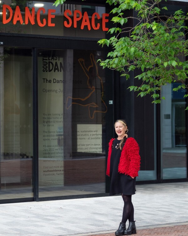 Cath James, Second Hand Dance Chair with long dark hair, wearing a black dress, bright red textured jacket, and black boots, standing outside The Dance Space in Brighton, with the studio’s glass facade and signage visible behind.