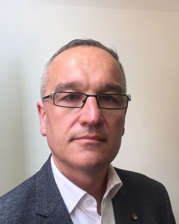 Jon Singleton, Second Hand Dance Board Member with short grey hair and glasses, wearing a white collared shirt and grey blazer, standing indoors against a plain light background.