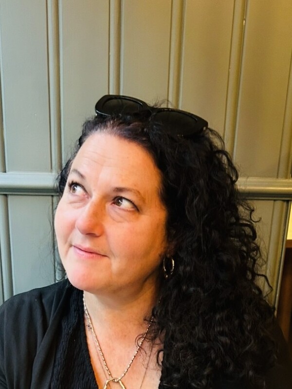 Louise Wildish, Second Hand Dance Board Member  Louise Wildish, Second Hand Dance Board Member with long curly dark hair, wearing a black top and sunglasses on her head, sits in front of a muted green paneled wall with a thoughtful expression.