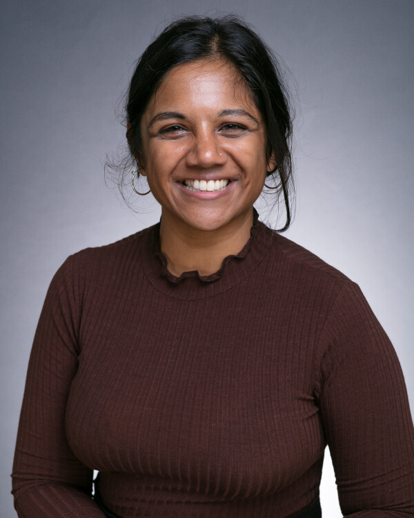 Rachna Joshi, Second Hand Dance Board Member Rachna Joshi, Second Hand Dance Board Member with dark hair tied back, wearing a brown ruffled top, smiles at the camera against a neutral gray background.