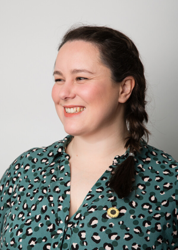 Rosie Heafford, Second Hand Dance Co-Director Rosie Heafford, Second Hand Dance Co-Director with dark braided hair, wearing a green patterned shirt with a sunflower pin, looking slightly to the left smiling against a plain light background.
