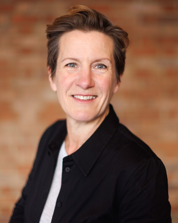 Claire Summerfield, Second Hand Dance Co-Director (Executive Producer), with short styled hair, wearing a black button-up shirt over a light top, facing the camera smiling against a warm blurred brick background.