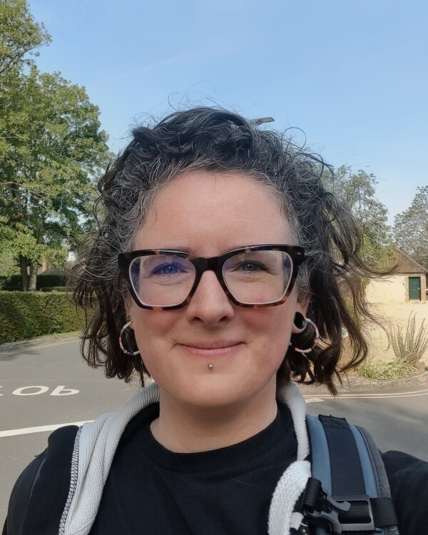 Emma Hayes, Second Hand Dance Head of Resources, with short curly hair and rectangular glasses, wearing a black top, facing the camera smiling against a bright leafy street background.